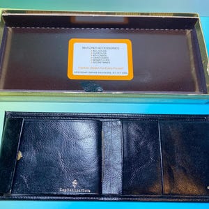 May include: An open black leather wallet with multiple compartments, displayed next to its original box. The box has a gold trim and a label listing "Matched Accessories" including billfolds and card cases. The wallet is embossed with "English Leather".