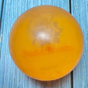 May include: An orange rubber ball with a faded illustration of a dog and the text "Buster Brown". The ball is a solid color and appears to be made of a soft material. The ball is sitting on a blue wooden surface.
