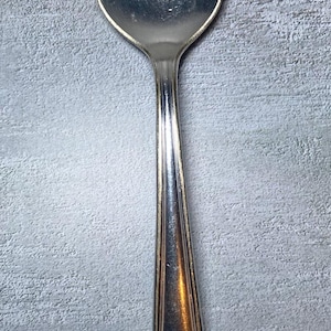 Op de afbeelding: Een zilveren lepel met een afgeronde kom en een decoratief handvat. Het handvat heeft een sierlijk bloemmotief en de initialen "H" en "K". De lepel staat tegen een grijze achtergrond met textuur.