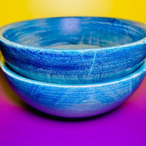 May include: Two stacked, round, blue wooden bowls. The bowls have a textured, painted finish with shades of light and dark blue. The bowls are on a purple surface with a yellow background.