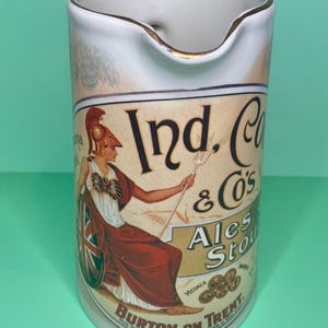 Puede incluir: Jarra de cerámica blanca con borde dorado y asa. La jarra presenta una etiqueta vintage con el texto "Ind. Co & Co's Ales Stout" y una ilustración de una mujer con toga sosteniendo un tridente.