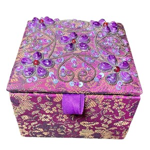 May include: A decorative purple jewelry box with a floral pattern and embellished with purple gems, sequins, and beads. The box has a matching purple ribbon closure and a gold floral pattern on the sides.