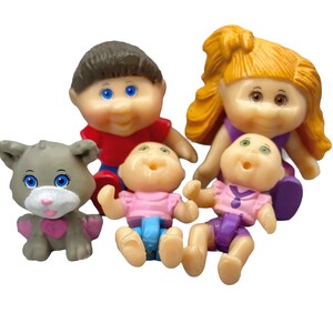 May include: A collection of five small plastic figurines including a gray cat with a pink heart, a child with brown hair in a red shirt, a larger child with orange hair in a purple outfit, and two infants in pink shirts with blue and purple bottoms.