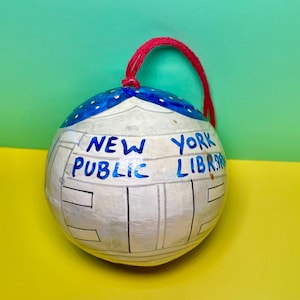 Puede incluir: Adorno redondo y blanco con una cuerda roja para colgar. La parte superior es azul con puntos blancos. Las palabras "NEW YORK PUBLIC LIBRARY" están escritas en azul. El adorno tiene un diseño de edificio.