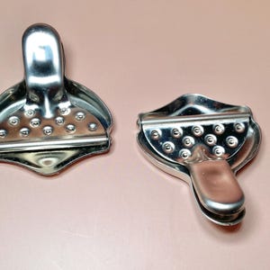 May include: Two stainless steel citrus juicers. Each juicer has a handle and a perforated bowl-shaped head. The juicers are designed to extract juice from lemons and limes. The juicers are shown on a light pink surface.