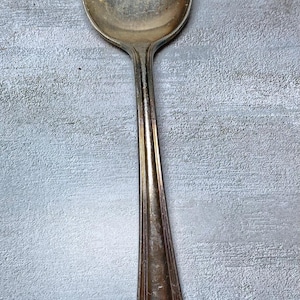 May include: A vintage silver spoon with a worn, reflective surface. The spoon has a long, slender handle with a decorative emblem near the end. The spoon is set against a textured, light gray background.