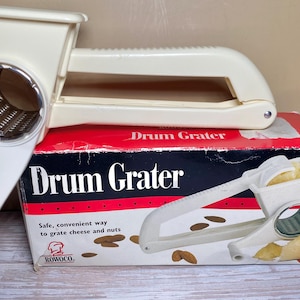 May include: Cream-colored drum grater with a handle, resting on a red, black, and white box. The box reads "Drum Grater" and features an illustration of the grater. The grater is designed for cheese and nuts.