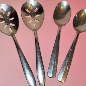 May include: Four stainless steel serving utensils on a pink background. Two slotted spoons with floral cutouts and two solid spoons with star and line designs on the handles. Kitchenware.