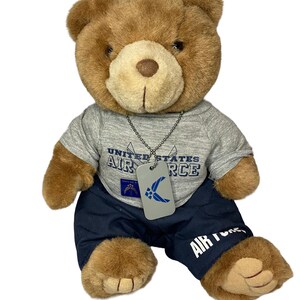 May include: A brown plush teddy bear wearing a heather gray t-shirt with the text United States Air Force and a blue emblem. The bear wears a silver-toned metal dog tag on a ball chain necklace and navy blue pants with the text Air Force printed.
