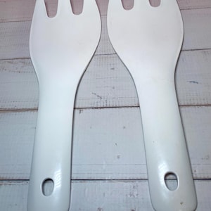 May include: Two white salad servers with a fork-like design. Each server has a long handle with a hole for hanging. The servers are made of a smooth, solid material and are designed for serving salads.