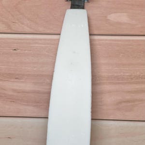 Op de afbeelding: Een keukengereedschap met een witte handgreep en een reeks kleine, cirkelvormige messen. De messen zijn verbonden met een metalen frame. De handgreep heeft een rechthoekige opening. De tool is waarschijnlijk om te snijden of te snijden.