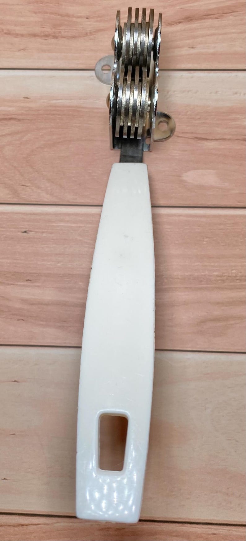 Op de afbeelding: Een keukengereedschap met een witte handgreep en een reeks kleine, cirkelvormige messen. De messen zijn verbonden met een metalen frame. De handgreep heeft een rechthoekige opening. De tool is waarschijnlijk om te snijden of te snijden.