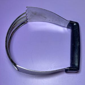 Può includere: Un frullatore per pasticceria in acciaio inossidabile con manico nero. Le lame curve sono collegate al manico, progettate per tagliare il burro nella farina. Lo strumento è posto su uno sfondo viola.
