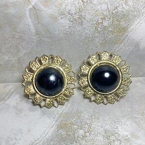 May include: A pair of vintage gold-tone earrings. Each earring features a large, glossy black center stone surrounded by a detailed, openwork gold-tone frame. The frames have a sunburst or floral design.