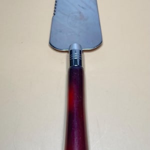 Könnte beinhalten: Ein Kuchenheber aus Edelstahl mit gezahntem Rand und burgunderfarbenem Griff. Der Griff hat eine tiefe, satte Farbe, die von einem dunklen Rot in einen helleren Farbton übergeht. Zum Schneiden und Servieren von Kuchen.