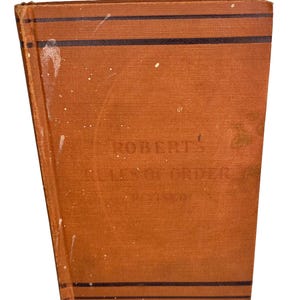 May include: A vintage hardcover book titled Robert's Rules of Order Revised with a textured orange fabric cover. The front cover features two horizontal black stripes at the top and bottom, with white paint splatters across the surface.