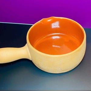 May include: A small, terracotta-colored ceramic pot with a handle. The pot is glazed on the inside and has a rough, textured exterior.