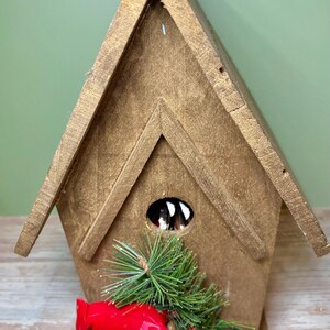 May include: A wooden birdhouse with a gold finish, featuring a decorative red cardinal and green pine sprigs. The birdhouse has a triangular roof and a circular opening. The birdhouse is a decorative item.