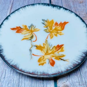Può includere: Un piattino in ceramica bianca con un bordo argentato e un disegno di tre foglie d'acero gialle e arancioni.