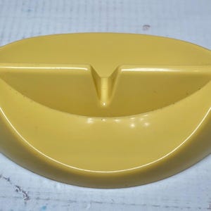 May include: Mustard yellow ashtray with a curved oval shape. The ashtray has a raised divider in the center and a small notch. The ashtray is made of a glossy material.