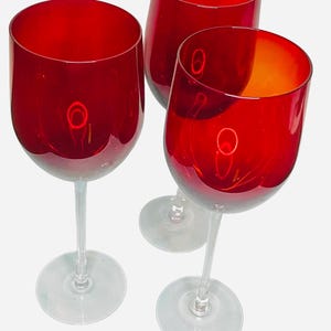 May include: Three wine glasses with deep red bowls and clear glass stems and bases. The glasses are arranged in a cluster, showing a smooth, rounded shape for each bowl. The red glass is translucent, allowing light to pass through the material.