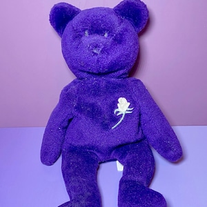 May include: A plush, purple teddy bear with a white rose embroidered on its chest. The bear has a classic design with a round head, small ears, and a soft, fuzzy texture. It is sitting on a purple surface.