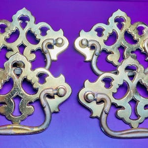 May include: Four antique brass drawer pulls with ornate, openwork designs. Each pull features a decorative backplate and a curved handle. The brass has a warm, aged patina, suggesting vintage or repurposed use. The background is a solid purple.