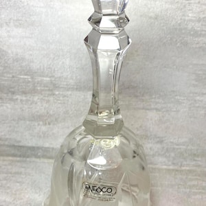 May include: A clear glass bell with a faceted top and a flared, scalloped base. The bell has a frosted floral design and a label that reads "Enesco". The bell is made of genuine lead crystal.