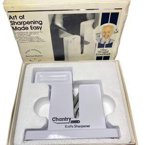 K&ouml;nnte beinhalten: Ein wei&szlig;er Chantry Messersch&auml;rfer mit V-f&ouml;rmigem Schlitz in einer wei&szlig;en Pappschachtel. Die Schachtel tr&auml;gt den Text Art of Sharpening Made Easy, The Frugal Gourmet und ein Portr&auml;t von Jeff Smith. Der Sch&auml;rfer liegt in einer wei&szlig;en Schaumstoffeinlage.