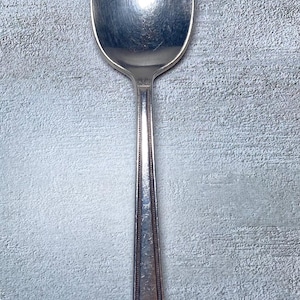 以下が含まれることがあります： 丸みを帯びたボウルと長く先細りのハンドルが付いた銀のスプーン。ハンドルには、一連の線と点で構成された装飾的なデザインがあります。スプーンは、テクスチャ加工された灰色の背景に配置されています。