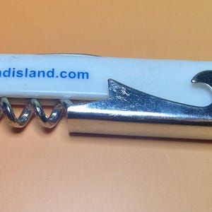 May include: A white and silver corkscrew with a bottle opener. The white handle has the text "www.baldheadisland.com" in blue. The corkscrew is made of metal and has a spiral design.