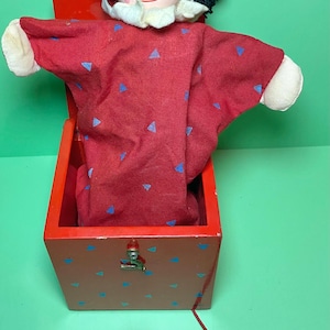 May include: A vintage Jack-in-the-box toy. The red wooden box features blue triangle accents. A red clown with a white ruffled collar and a red hat pops out. The clown has black hair, a red nose, and a smiling face.