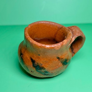 May include: A handmade, rustic brown ceramic mug with a handle. The mug has a rounded body and a slightly irregular rim. Dark green and black markings add character to the exterior. The mug is likely suitable for hot beverages.