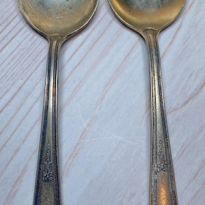 May include: Two antique silver spoons with round bowls and ornate, detailed handles. The spoons have a tarnished, aged appearance, suggesting they are vintage or heirloom pieces. The handles feature intricate floral and scrollwork designs.