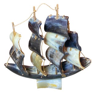 May include: A decorative model sailboat with a dark hull and sails made from a mix of light and dark horn. The sails are attached to wooden masts with twine rigging. The boat sits on a rectangular base.