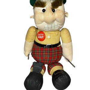 May include: A plush doll dressed as a golfer wearing a tan hat, a yellow collared shirt with a red button reading #*?*! GOLF!, a black belt, and red plaid pants. The doll has thick brown eyebrows, a gritted teeth expression, and black shoes.
