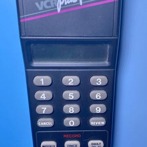May include: A black VCR Plus device with a keypad featuring numbers and function buttons. The device has the text "VCR Plus" and "GEMSTAR" printed on it. The device is designed for programming VCRs.