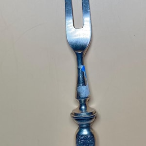 May include: Silver-toned carving fork with two tines. The handle features an ornate design with a grape cluster and bird motif. The fork is approximately 10 inches long.