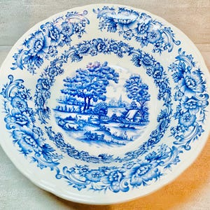May include: A white ceramic serving bowl with a blue floral pattern and a central landscape scene. The bowl features a scalloped edge and is decorated with detailed blue floral designs.