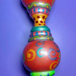 May include: A decorative, hand-painted object with a red, bulbous top and bottom, connected by a narrower, colorful section. The red sections feature swirling, colorful designs. The base is silver-colored. The background is purple.