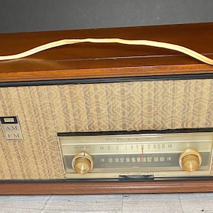Puede incluir: Consola de radio Philco AM/FM vintage en un mueble de madera. La radio tiene una cubierta de altavoz tejida de color claro y un dial con perillas doradas. El mueble es de color marrón oscuro con paneles laterales decorativos y un cable de alimentación.
