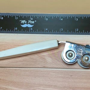 Op de afbeelding: Een vintage blikopener met een witte handgreep en een zilverkleurig snijmechanisme met dubbele wielen. Een zwarte liniaal met witte markeringen en de tekst "Mr. Pen" staat op de achtergrond. De blikopener ligt op een houten oppervlak.