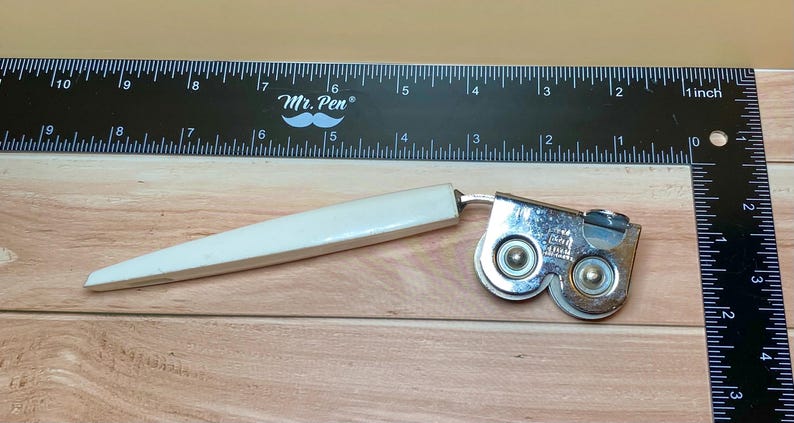 Op de afbeelding: Een vintage blikopener met een witte handgreep en een zilverkleurig snijmechanisme met dubbele wielen. Een zwarte liniaal met witte markeringen en de tekst "Mr. Pen" staat op de achtergrond. De blikopener ligt op een houten oppervlak.