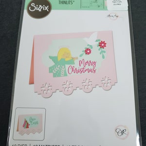 Op de afbeelding: Een roze kaart met een gestanst ontwerp met een engel met een halo en vleugels, die een trompet vasthoudt. De kaart zegt "Merry Christmas" en heeft een witte duif en groene bladeren. De kaart is verpakt met 12 stansen.