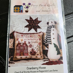 Può includere: Uno schema di ricamo a punto croce per una casa di mirtilli rossi, parte di una serie chiamata "Case su Peppermint Lane". Lo schema è stato progettato da Lori Pengelly ed è il grafico 5 di 9 della serie. Lo schema presenta una casa rossa con finiture bianche e una staccionata bianca. C'è un pupazzo di neve con una sciarpa a strisce bianche e nere e un cappello verde a destra della casa.