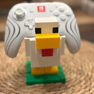 May include: A white, blocky, pixel-style chicken figure serving as a controller stand. The chicken has a yellow beak, a red wattle, black eyes, and yellow feet on a green base. A white wireless game controller with gray swirl patterns rests behind it.