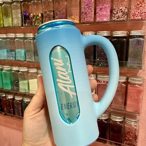 Puede incluir: Un enfriador de latas azul claro con asa, que sostiene una lata de bebida energ&eacute;tica Alani. El enfriador tiene un recorte para mostrar la etiqueta de la lata. El fondo presenta estantes llenos de peque&ntilde;os frascos de purpurina colorida.