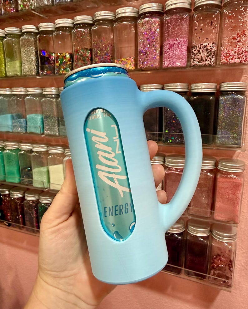 Puede incluir: Un enfriador de latas azul claro con asa, que sostiene una lata de bebida energ&eacute;tica Alani. El enfriador tiene un recorte para mostrar la etiqueta de la lata. El fondo presenta estantes llenos de peque&ntilde;os frascos de purpurina colorida.