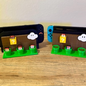 May include: Two video game console docks featuring a brown brick wall design with a yellow question mark block, a white cloud, and three green pipes. Each dock holds a handheld console and several small game cartridges in the pipe and block slots.