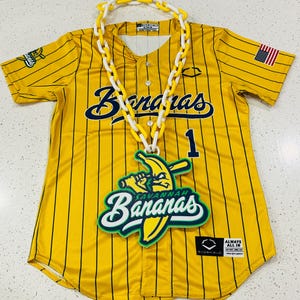 May include: Yellow baseball jersey with black vertical stripes, the word "Bananas" in navy blue script, and the number 1. A white and yellow chain necklace and a green and white banana logo are on the jersey. An American flag patch is on the sleeve.